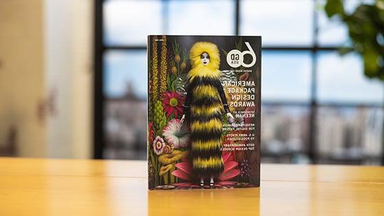 Graphic 设计 USA’s April 2023 issue sits upright on a wooden table. The cover features a female doll wearing a long furry black-and-yellow dress with a hood. She is standing in front of a psychedelic floral background.