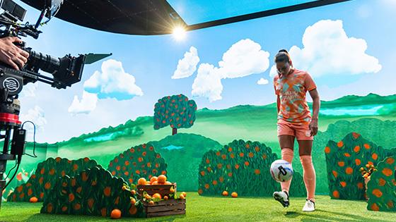 An Orlando Pride player kicks a soccer ball toward a camera on a film set that features an animated orange grove.