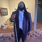 Andre Woods stands outdoors in front of a restaurant. 他穿着一套蓝色的西装，戴着太阳镜. 他拄着一根手杖.