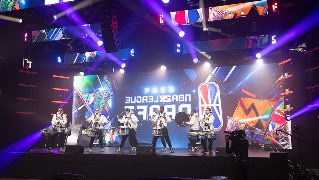 A drumline is performing on a stage in front of a large LED screen that reads “NBA 2 k 联盟 Draft.”