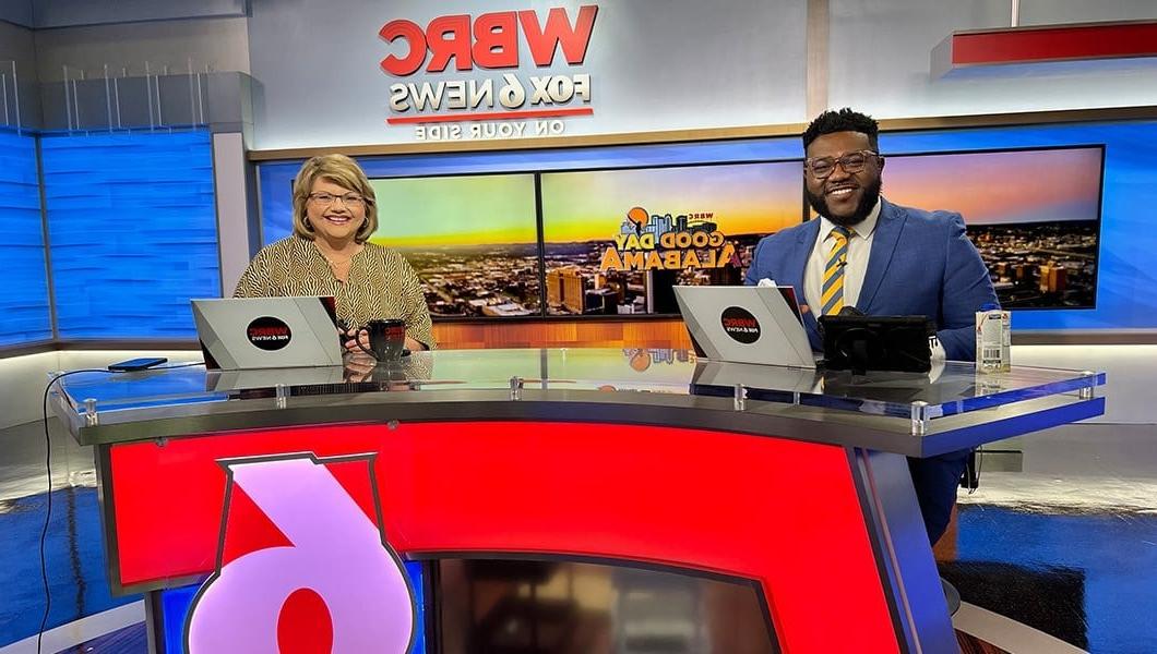 Toi Thornton sits at a news desk with his co-anchor. A screen behind them shows the Birmingham skyline and the Good Day Alabama logo.